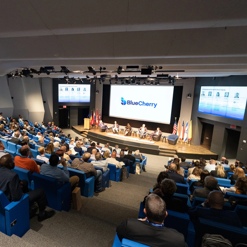 Auditorium setting with people in rows looking at panel on stage, BlueCherry on screen
